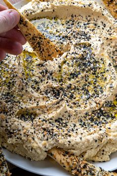 a hand dipping hummus into a white bowl filled with mustard and black sesame seeds