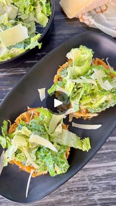 two black plates filled with lettuce, cheese and other food on top of a wooden table