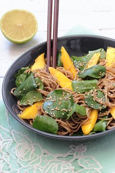 a black bowl filled with noodles, mangos and greens next to chopsticks