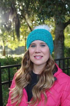 a woman wearing a pink jacket and a blue knitted beanie smiles at the camera