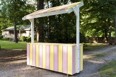 a yellow and pink striped ice cream stand in the middle of a park with lots of trees