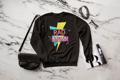 "This totally tubular Rad Mom sweatshirt is perfect for moms who love the 80s or 90s. The retro graphic style and Rad Mom text makes a perfect gift for mother's day or mom's birthday! We also offer this design on a t-shirt and classic coffee mug. Also available for Dads! See all our \"rad\" themed items: https://www.etsy.com/shop/LifeLikeDesignsShop?search_query=rad This super soft sweatshirt has a classic fit for a comfortable feel. Unisex sizing for great versatility. .: 50% Cotton 50% Polyest Retro Cotton Sweatshirt With Funny Print, Trendy Cotton Sweatshirt For Mother's Day, Black Graphic Print Sweatshirt For Mother's Day, Retro Cotton Sweatshirt With Graphic Print, Black Graphic Print Mother's Day Sweatshirt, Mother's Day Black Graphic Print Sweatshirt, 80s Mom, Mom Texts, 90s Mom