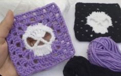 a hand is holding a crocheted square with a skull on it and two skeins of yarn next to it