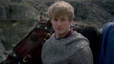 a young man in armor standing next to a brown horse on a rocky mountain side