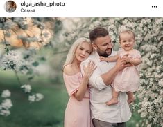 a man and woman holding a baby in front of some white blossomy trees with their arms around each other