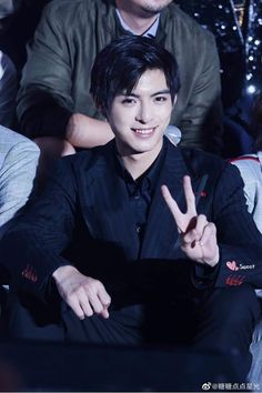 a man in a suit sitting down and making the peace sign with his hand while people watch from behind him