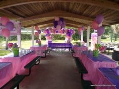 an outdoor picnic area with purple and pink tablecloths, balloons and flowers on the tables