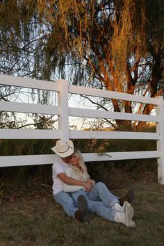 a man and woman sitting on the ground next to a white fence with trees in the background