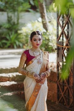 a woman in a white and gold sari with jewelry on her neck, standing outside