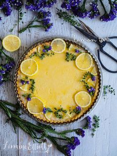 a pie with lemons and purple flowers around it on a white wooden table next to scissors