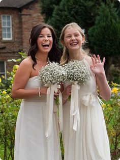 two women in white dresses standing next to each other with flowers on their hands and smiling at the camera