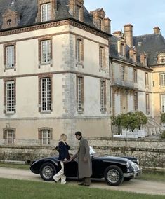 two people standing next to an old fashioned car in front of a large stone building