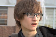 a young man with glasses and a backpack on his shoulder looking at the camera while standing in front of a building