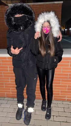 two people standing next to each other in front of a brick wall wearing winter clothes