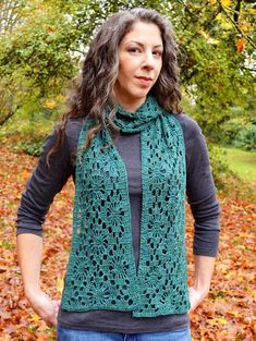 a woman wearing a green crochet scarf standing in front of trees with leaves on the ground