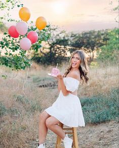 a woman is sitting on a stool holding a cake and balloons in the air as she poses for a photo