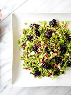 a white plate topped with greens and blackberries next to a wooden utensil