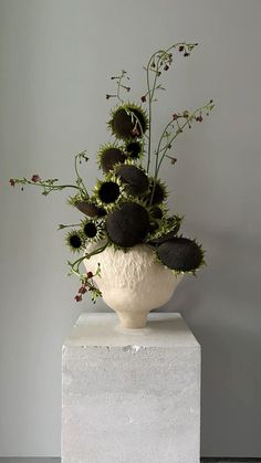 a white vase filled with lots of flowers on top of a marble block in front of a gray wall