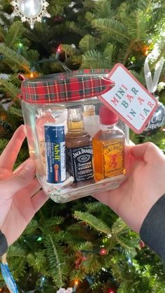 someone holding up a small jar filled with different types of alcohol in front of a christmas tree