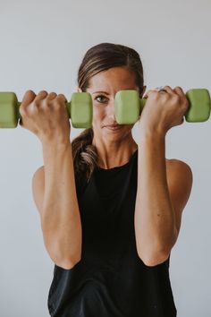 a woman lifting two green dumbbells in front of her face and looking at the camera