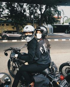 two people sitting on a motorcycle wearing helmets and protective gear, with cars in the background