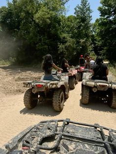 four people riding on four wheelers in the sand with trees and bushes behind them