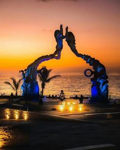 an artistic sculpture in front of the ocean at sunset with people standing on it and one person doing a handstand