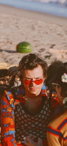 two people sitting on the beach looking at their cell phones and one person wearing red sunglasses