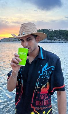 a man wearing a hat and holding a cup in his hand while standing on the beach