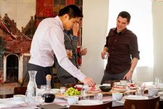 two men standing over a table with food on it and wine glasses in front of them