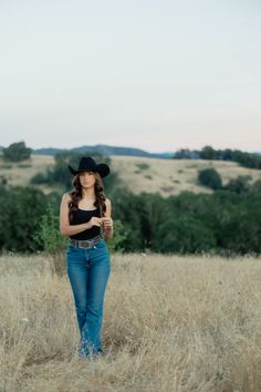 a woman standing in the middle of a field wearing a black cowboy hat and jeans