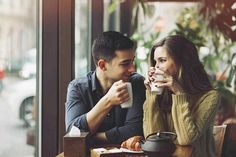 a man and woman sitting at a table with cups in their hands, looking into each other's eyes