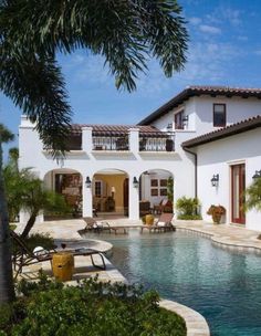 an outdoor swimming pool in front of a white house with palm trees and chairs around it