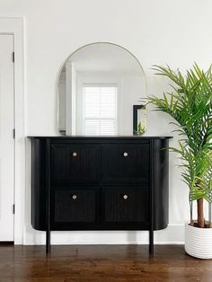 a black dresser sitting next to a mirror on top of a wooden floor near a potted plant