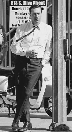 black and white photograph of a man leaning against a pole with his hands in his pockets