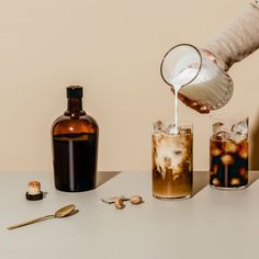 a person pouring something into two glasses on a table with bottles and spoons next to them