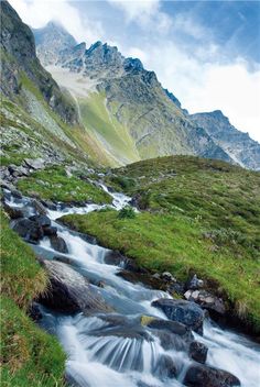 a small stream running through a lush green mountain valley with tall mountains in the background