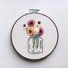 an embroidery project with flowers in a mason jar on a white surface, hanging from a hoop