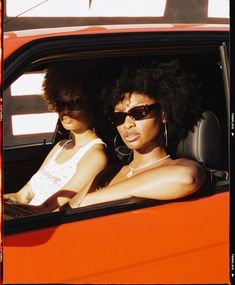 two women sitting in the drivers seat of an orange car, both wearing sun glasses