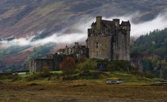 an old castle in the middle of a mountain range with fog coming off it's sides