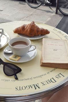 a table topped with a cup of coffee next to a croissant and book