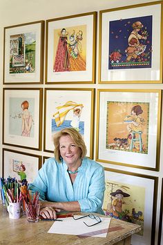 a woman sitting at a desk with many pictures on the wall behind her and colored pencils in front of her