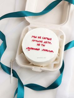 a white cake with red writing on it in a box next to a blue ribbon