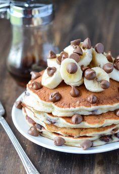 a stack of pancakes topped with banana slices and chocolate chips on a white plate next to a fork