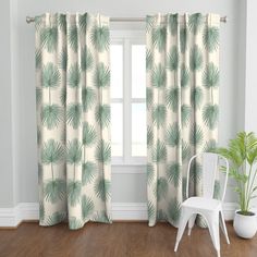 a white chair sitting next to a window covered in green palm leaf print curtain panels