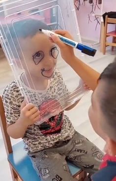 a little boy sitting in a chair with a fake face on his head and an adult brushing it's hair