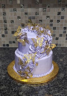 a three tiered cake with purple flowers and gold butterflies on the top is sitting on a marble countertop