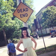 a woman in a graduation gown standing under a dead end sign on the side of a road