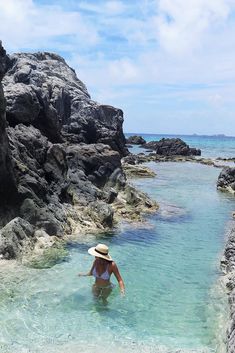 a woman wading in the clear blue water near some rocks and sand, with a hat on her head