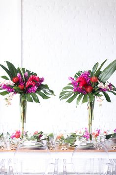 two vases filled with flowers and greenery on top of a table covered in wine glasses